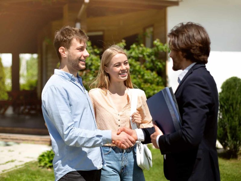 Young married couple shaking hands with realtor, making deal about house purchase outside